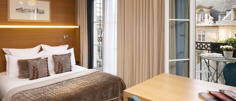 Quarto de hotel com cama, cabeceira de madeira, mesa com cadeiras e vista para prédios pela janela.