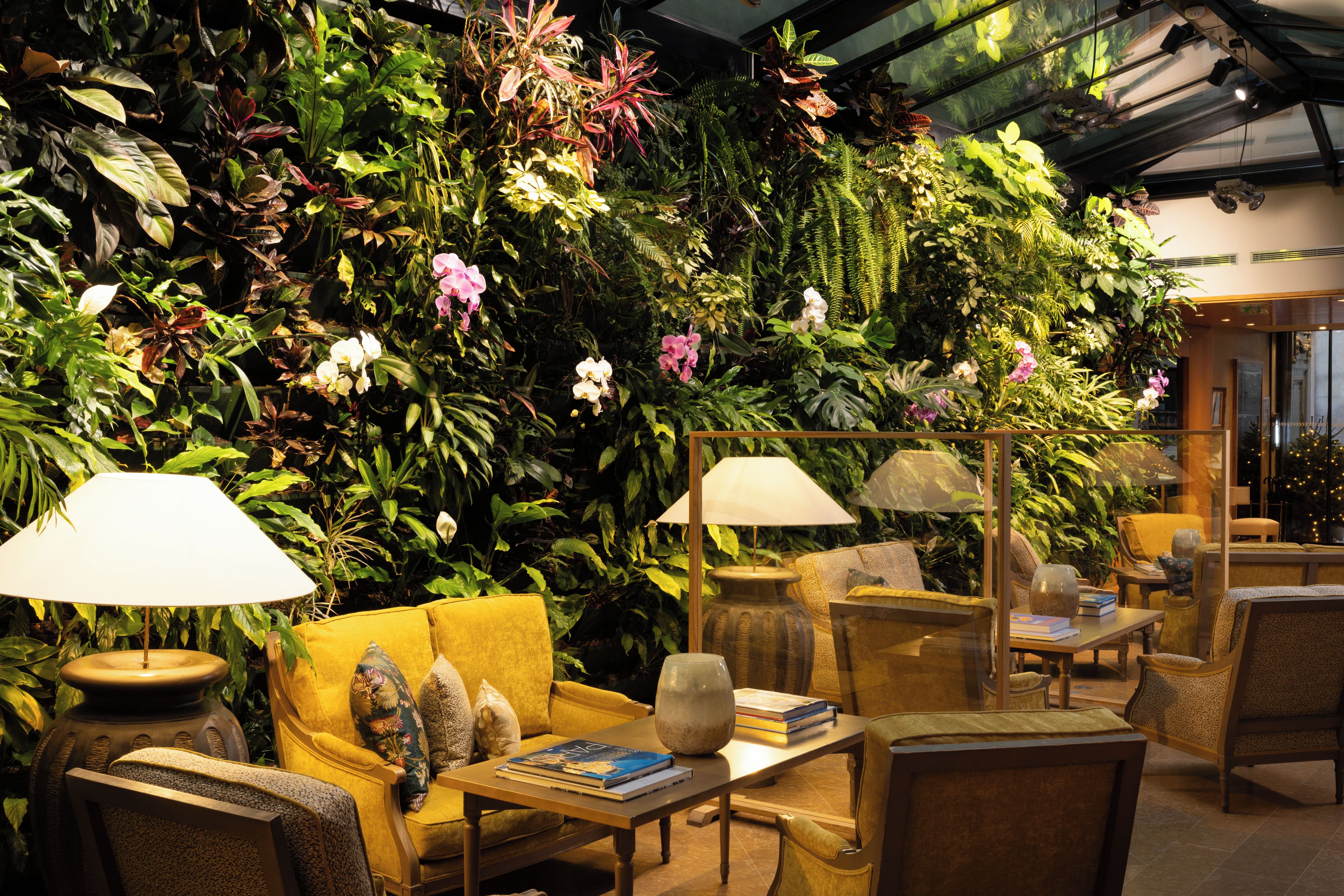 A room with a large green wall of plants, yellow sofas, and wooden tables with lamps.