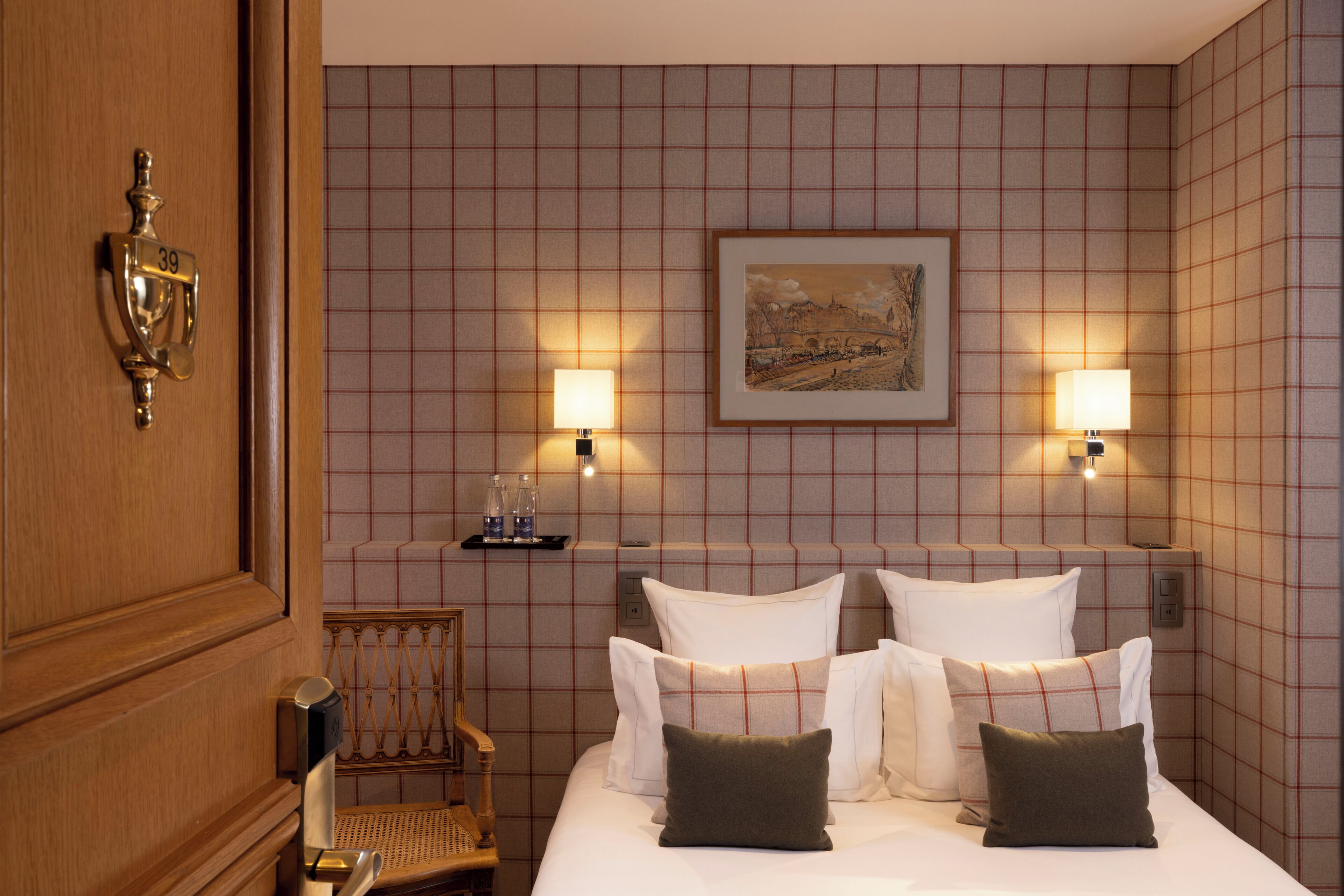 A hotel room with a bed, two wall-mounted lamps, a framed picture, a small shelf with bottles, and a wooden chair.