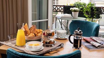 Una bandeja de desayuno con croissants, un vaso de zumo de naranja, un bol de cereales y una cafetera sobre una mesa con una silla.