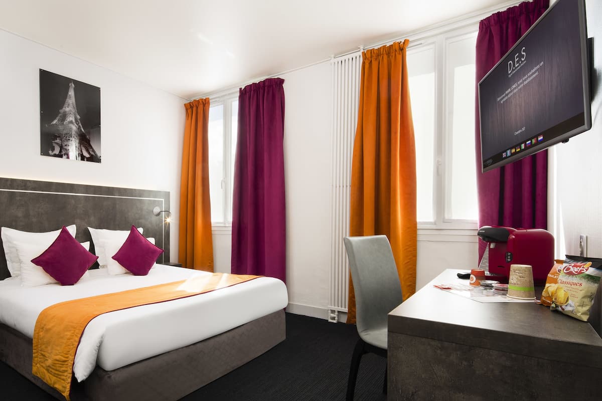 Una habitación de hotel con cama, escritorio, silla, televisión y ventana con cortinas.