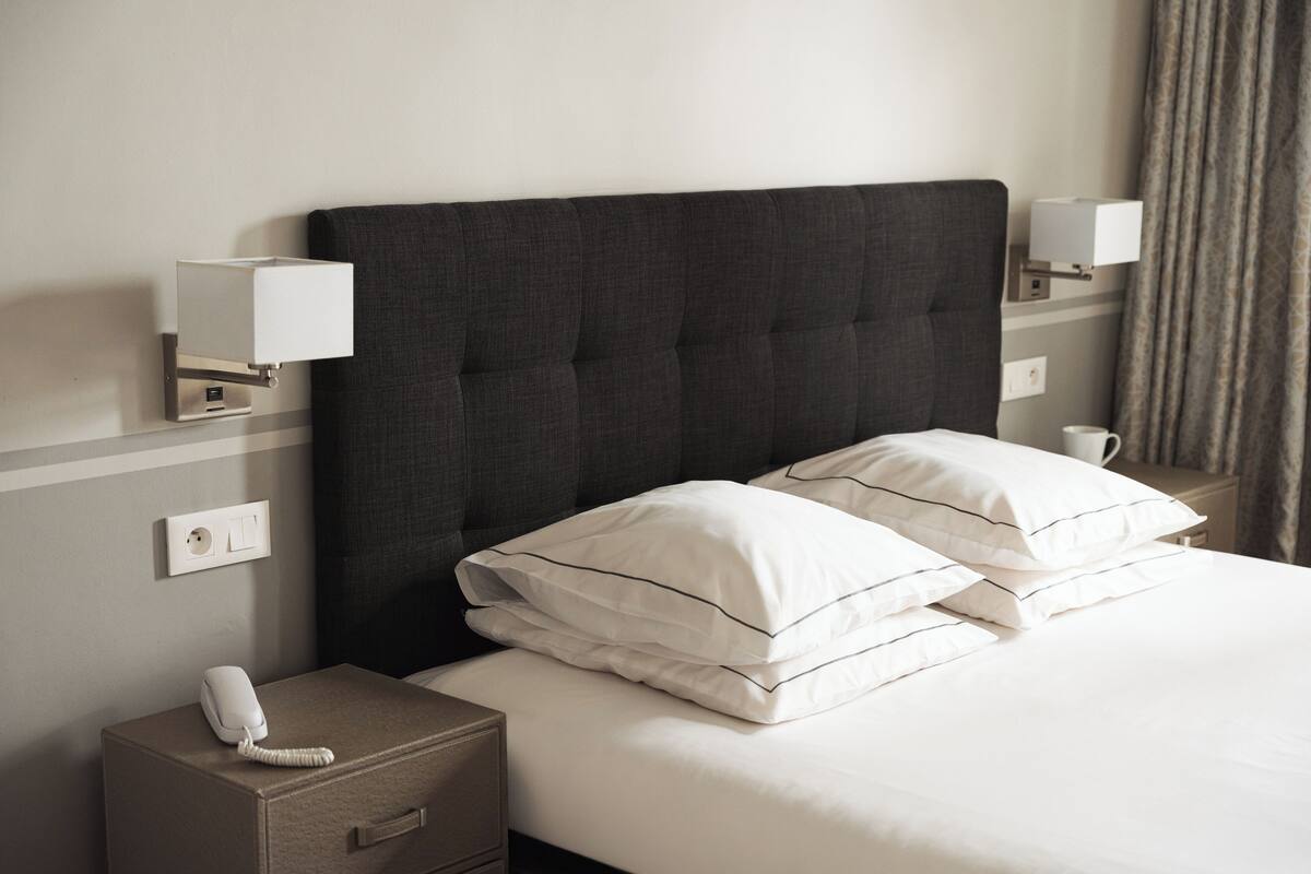 A neatly made bed with white linens and two pillows, a bedside telephone, and a wall-mounted lamp on a nightstand.