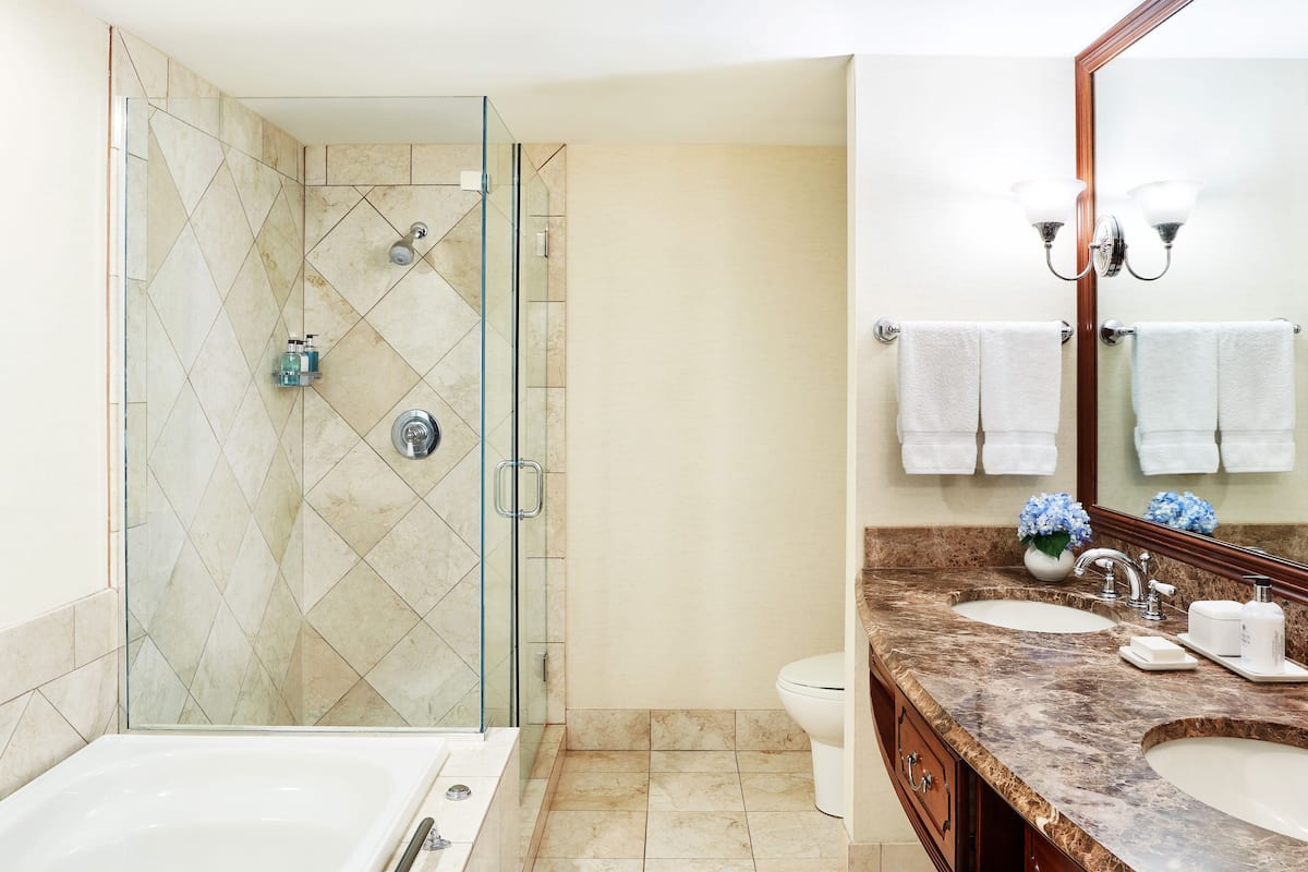 A bathroom with a glass-enclosed shower, a bathtub, two sinks, and a large mirror with wall-mounted lights.