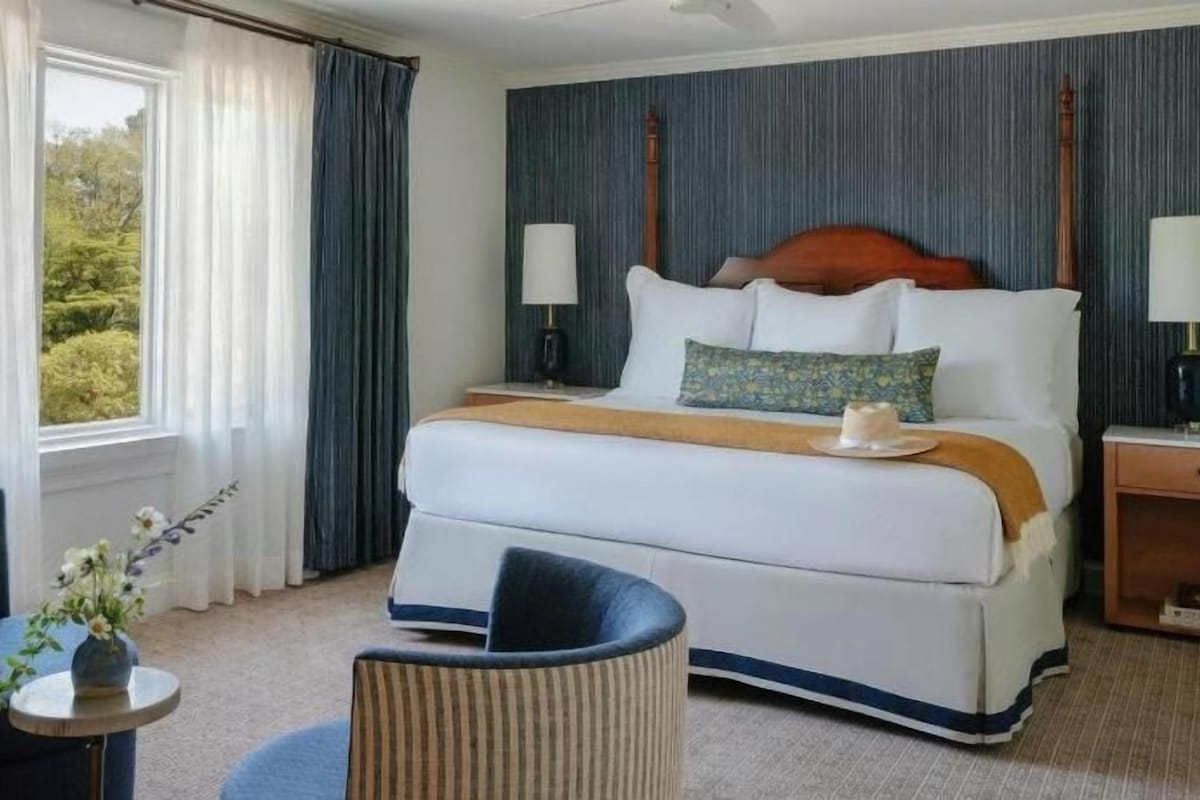 A hotel room with a large bed, two bedside tables, a round coffee table, and a view of the outdoors.