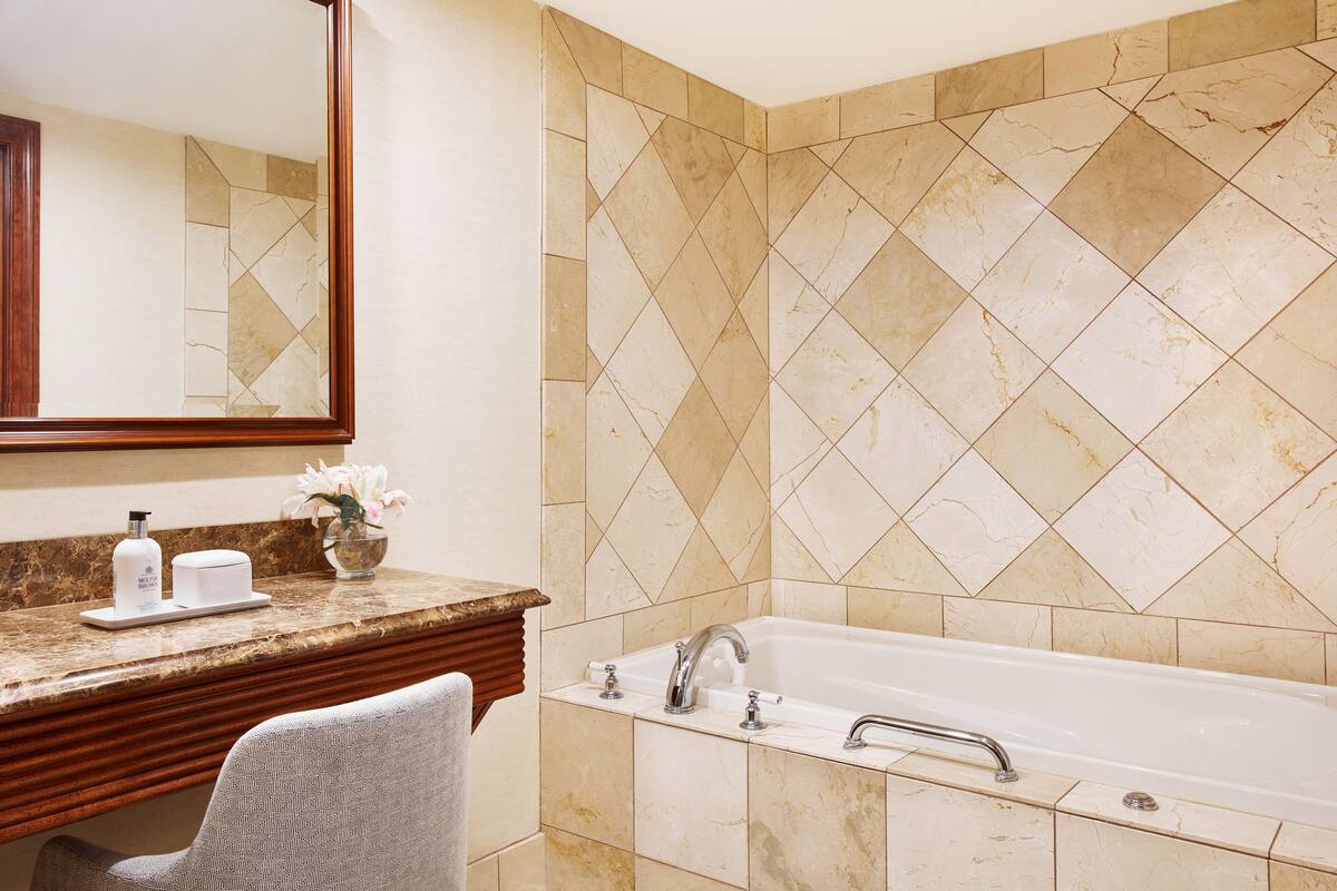 A bathroom with a marble-tiled wall, a bathtub, a vanity with a mirror, and a chair.