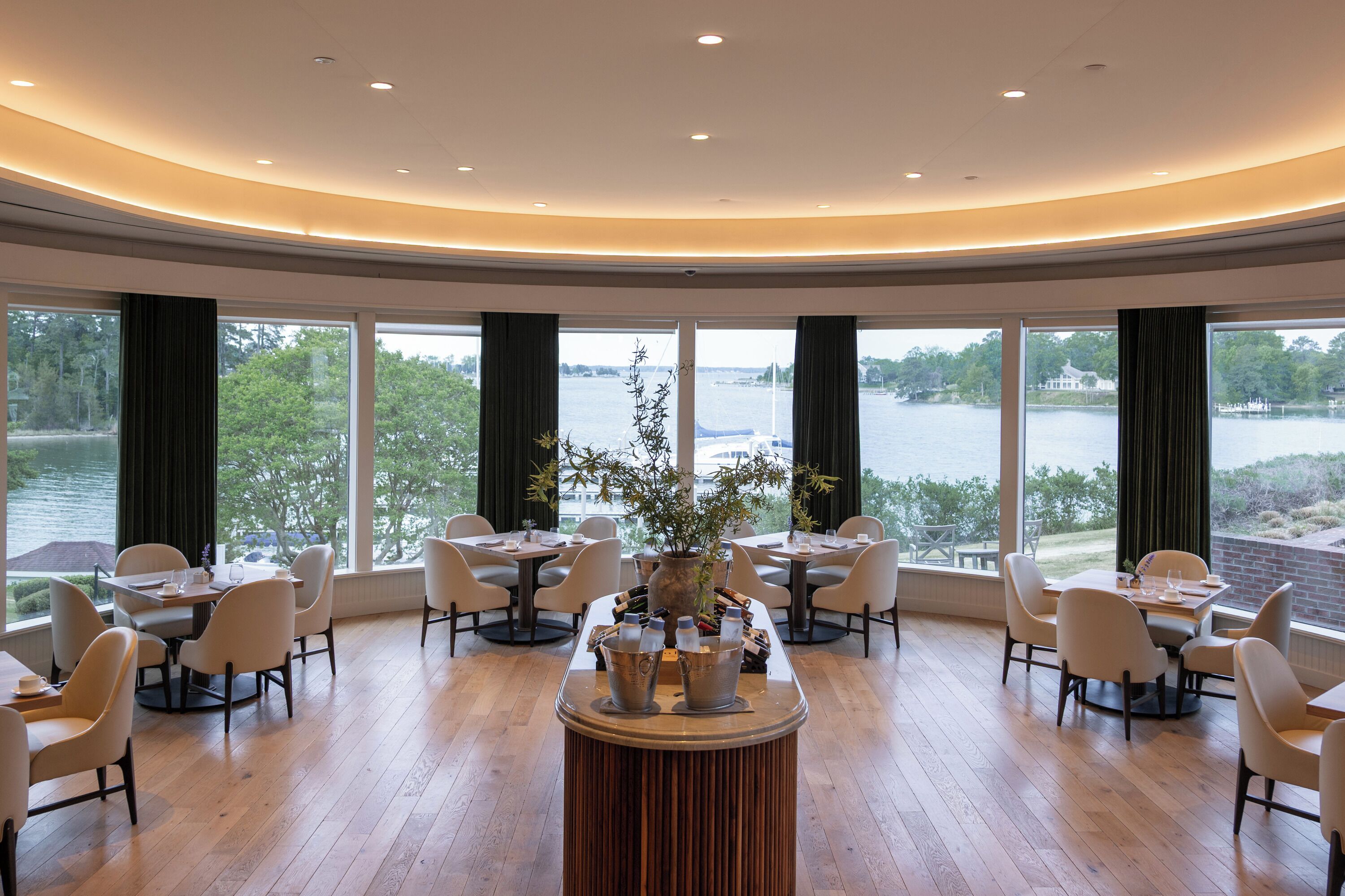 A spacious dining area with multiple tables and chairs, large windows offering a view of a lake and trees, and a central decorative plant arrangement.