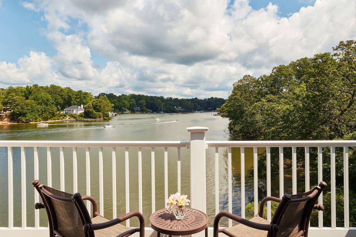 A balcony with two chairs and a table, overlooking a river with boats and houses in the background.