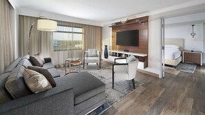 A modern hotel room with a grey sofa, a dining table, and a TV mounted on the wall.