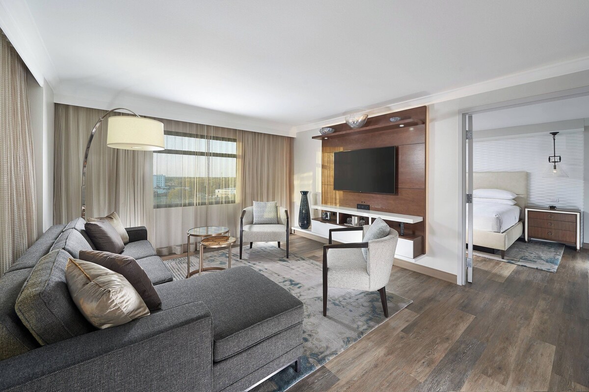 A modern hotel room with a grey sofa, a dining table, and a TV mounted on the wall.