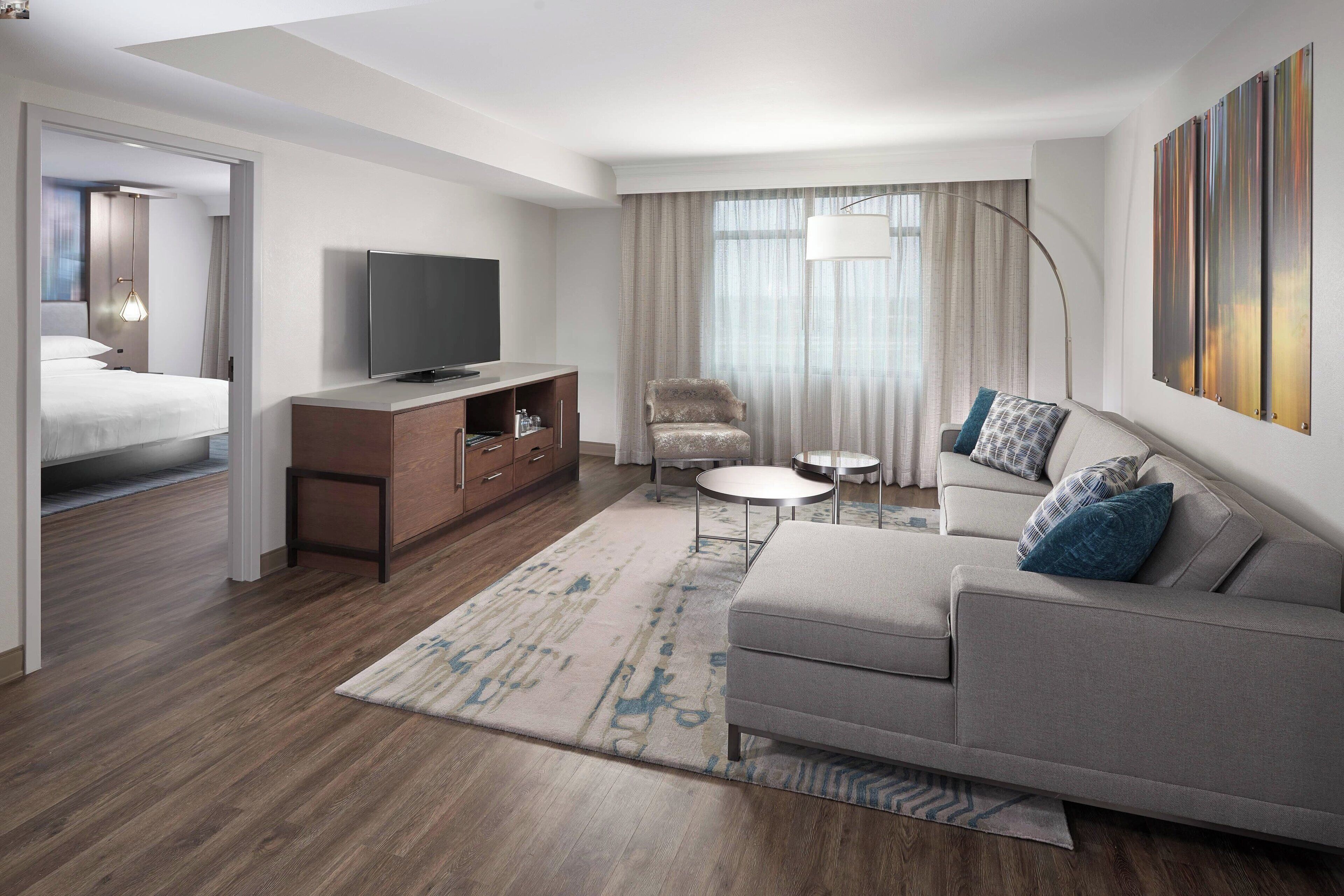Habitación de hotel moderna con sofá, televisión, sillón, una mesita redonda y un gran ventanal con cortinas.