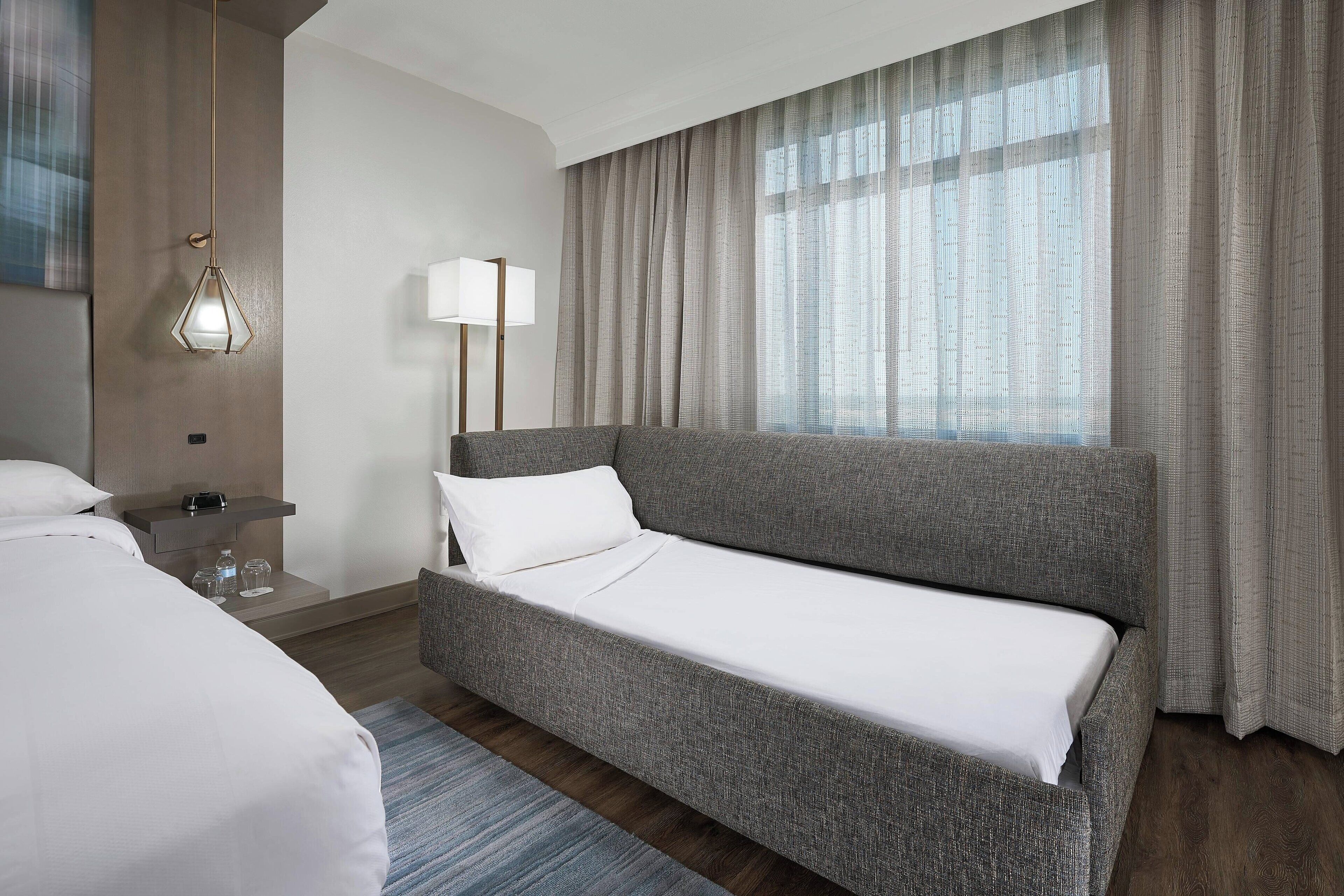 Una habitación de hotel moderna con un sofá cama gris, una mesita de noche con lámpara y un amplio ventanal con cortinas.