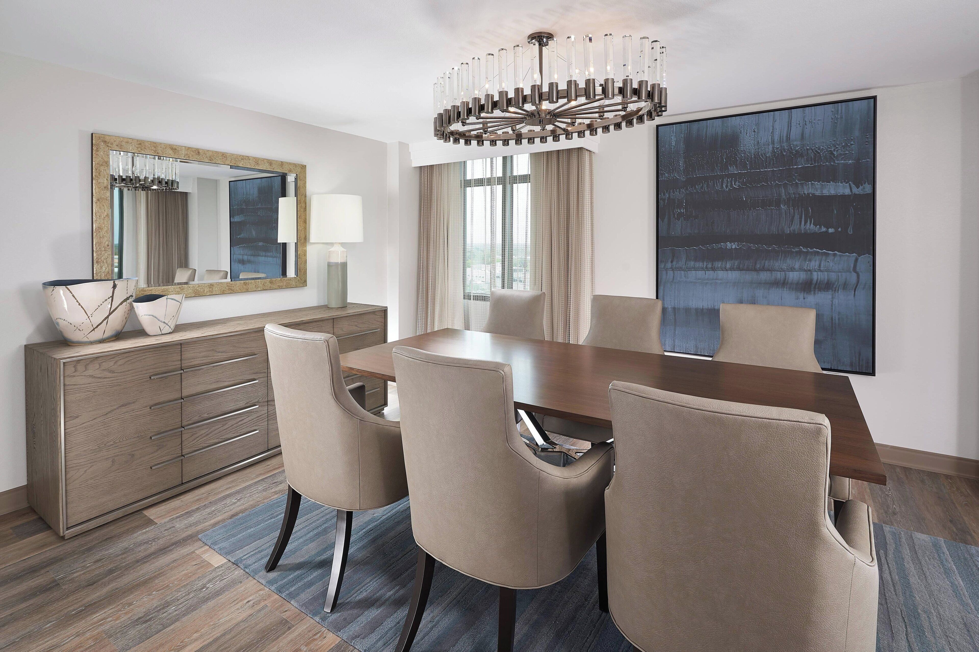 Un comedor moderno con una mesa de madera, sillas color beige, un espejo grande y una araña.