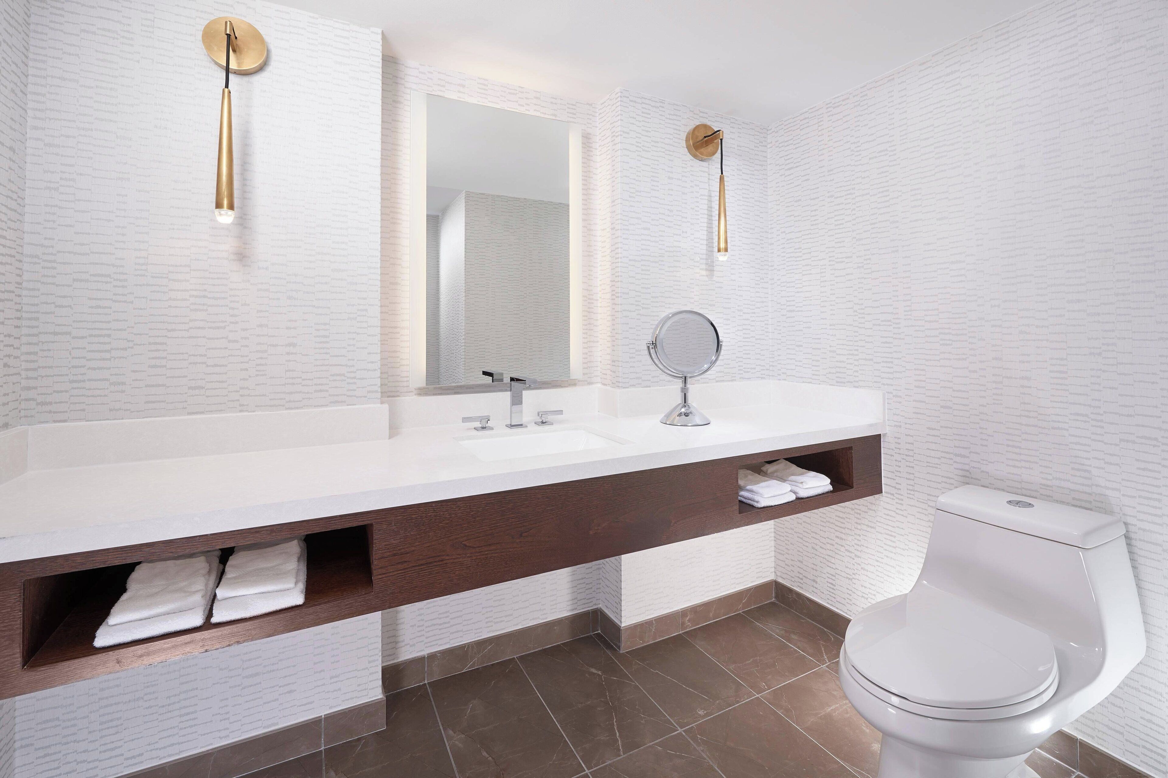 A modern bathroom with a white brick wall, a white sink with a wooden vanity, a round mirror, and a white toilet.
