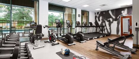 Un gimnasio con diverso equipo de ejercicio, amplias ventanas y vistas al exterior con vegetación.