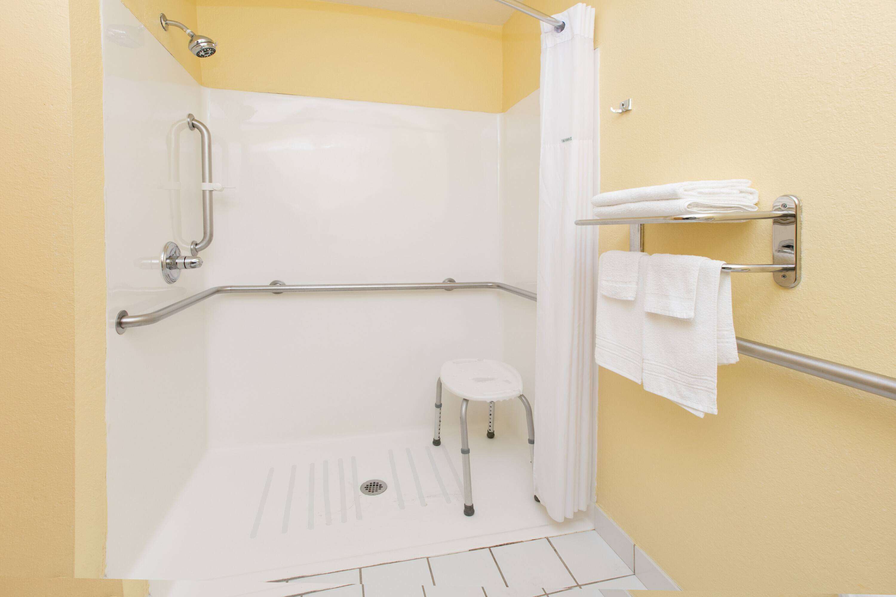 A wheelchair-accessible shower with grab bars, a step stool, and a towel rack with folded towels.