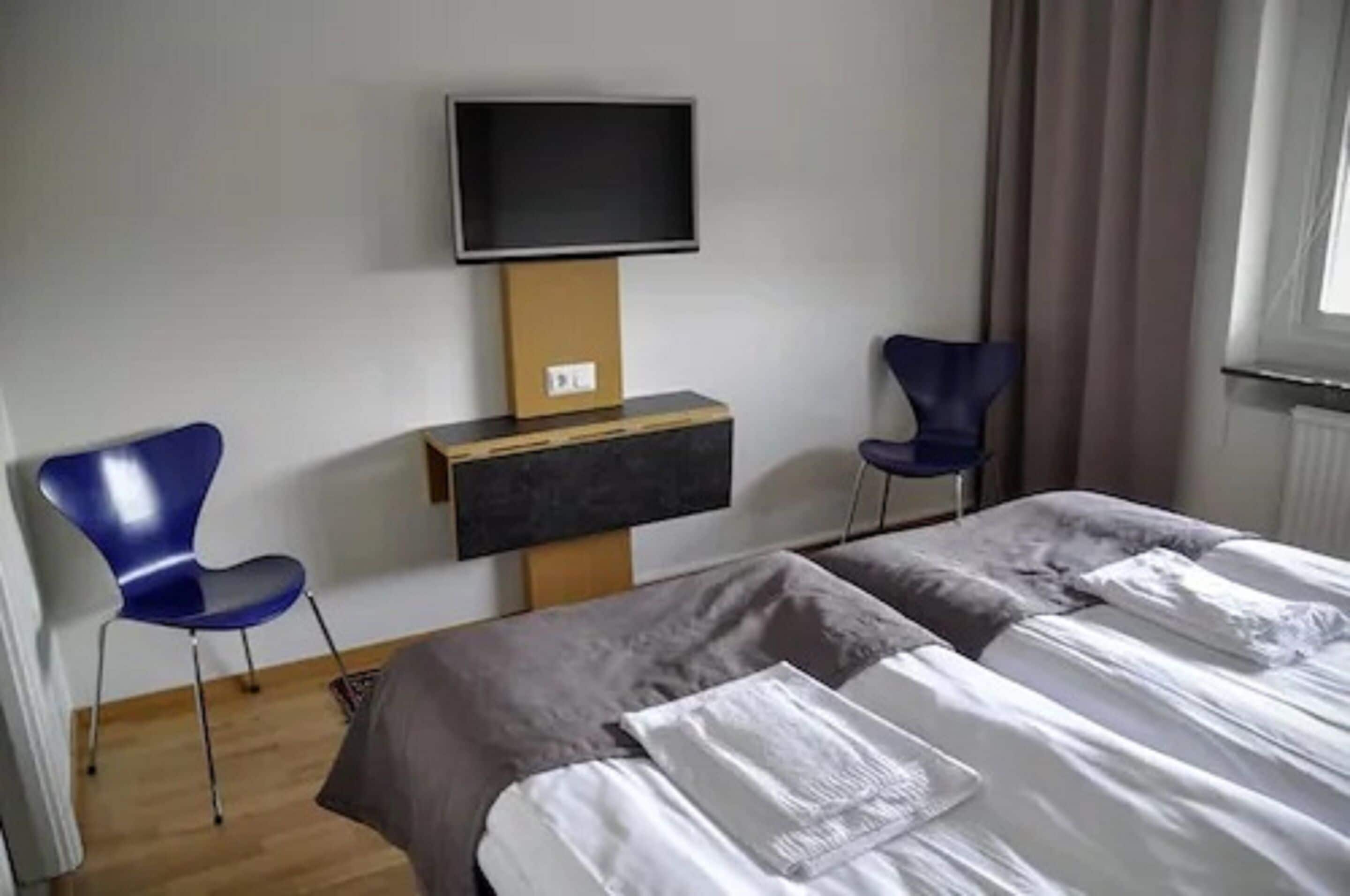 Une chambre d’hôtel avec deux lits, un téléviseur fixé au mur et deux chaises bleues.