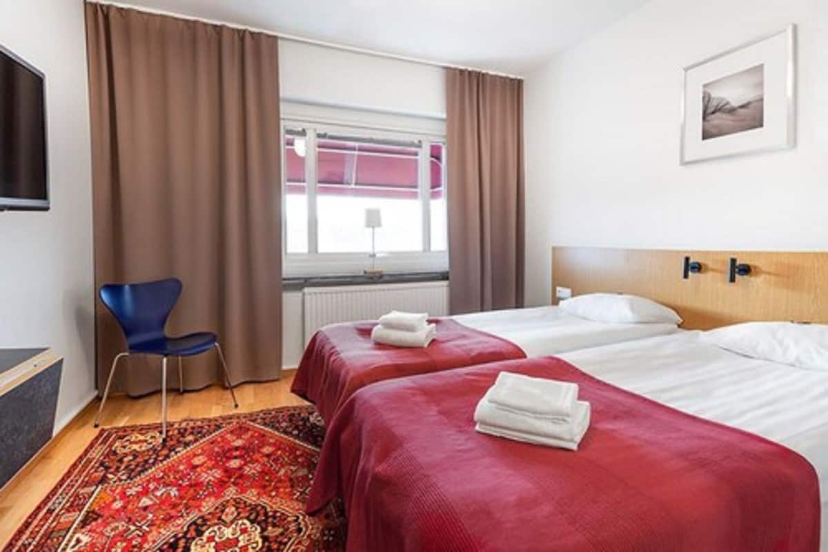 A hotel room with two beds, a red carpet, a blue chair, and a window with curtains.