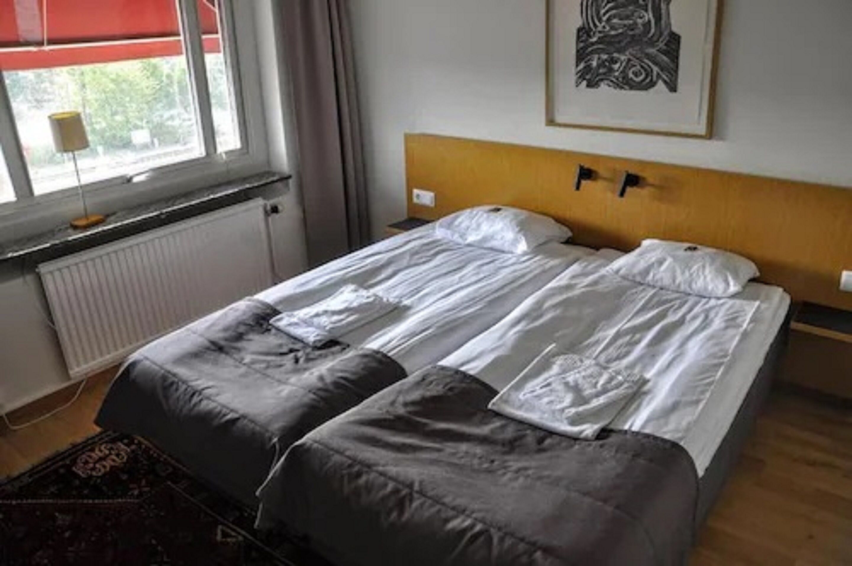 Een tweepersoonsbed met houten hoofdeinde, twee nachtkastjes, een radiator, een raam met rode jaloezieën en een lijstwerk met kunst erop.