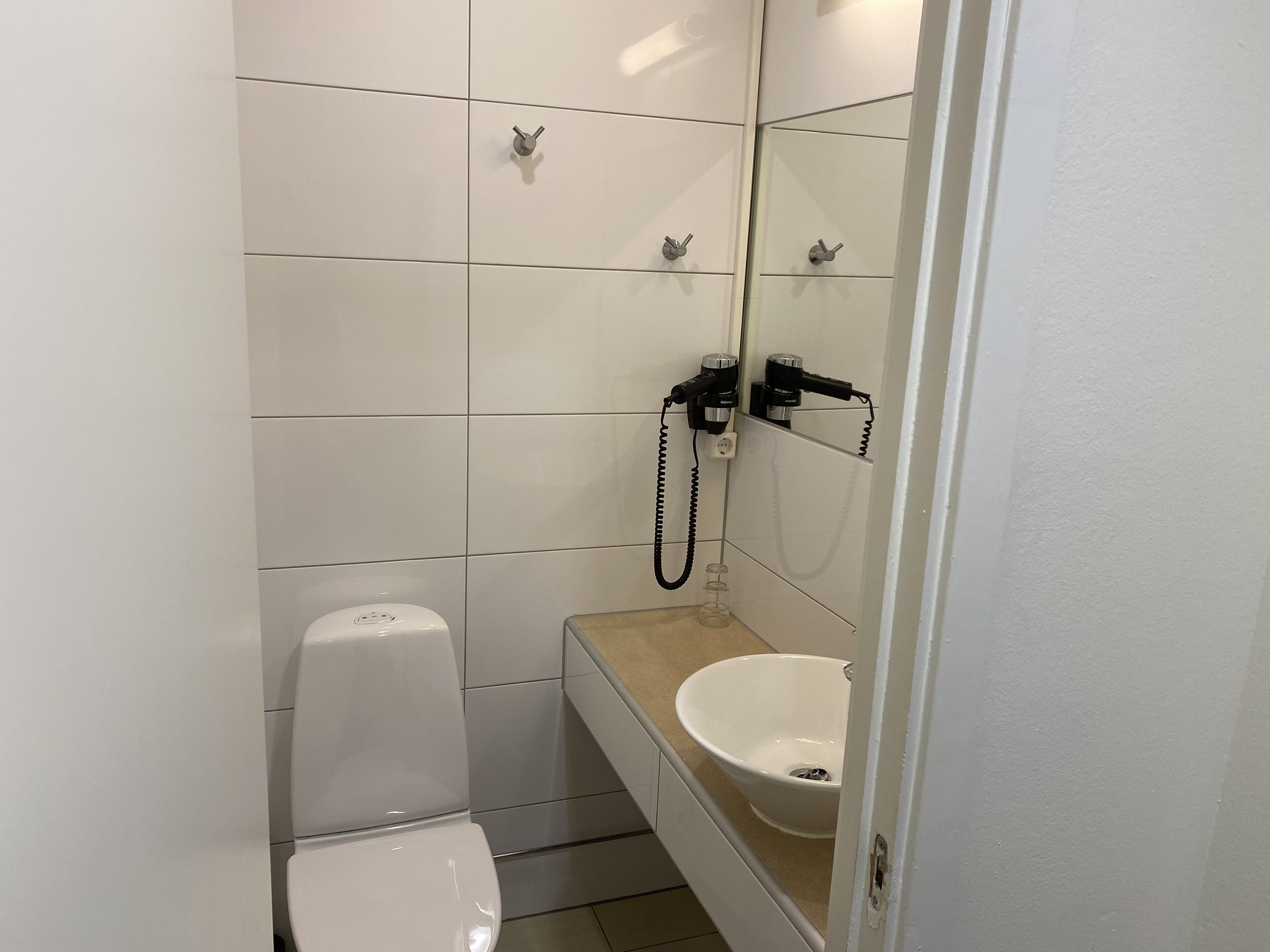 Une petite salle de bain compacte, avec des carreaux blancs, des toilettes, un lavabo avec un miroir et une douchette à main.