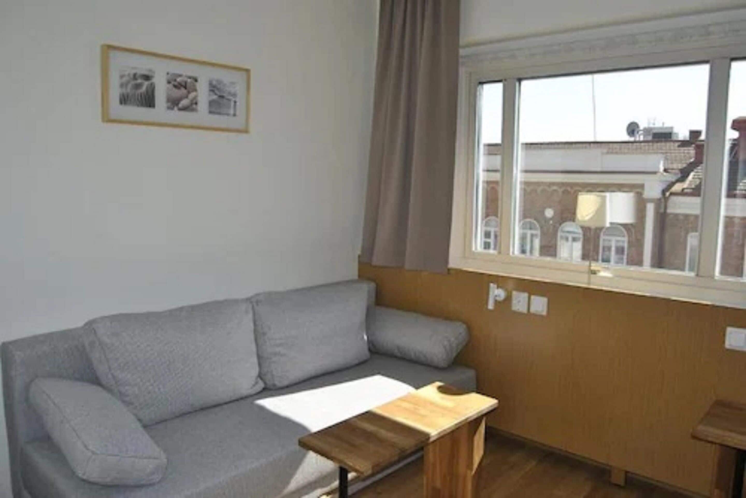 A room with a grey sofa, a wooden side table, a window with curtains, and a framed picture on the wall.