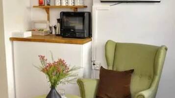 A compact living space with a kitchenette, a green armchair, a small round table with a vase, and a flat-screen TV mounted on the wall.