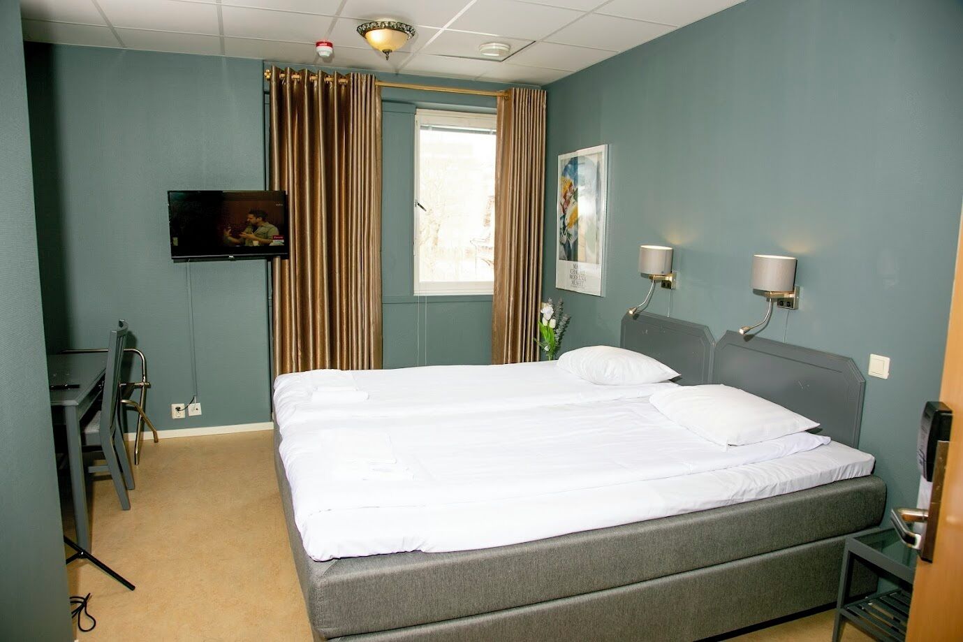 A hotel room with a large bed, a desk, a chair, a TV, and a window with curtains.