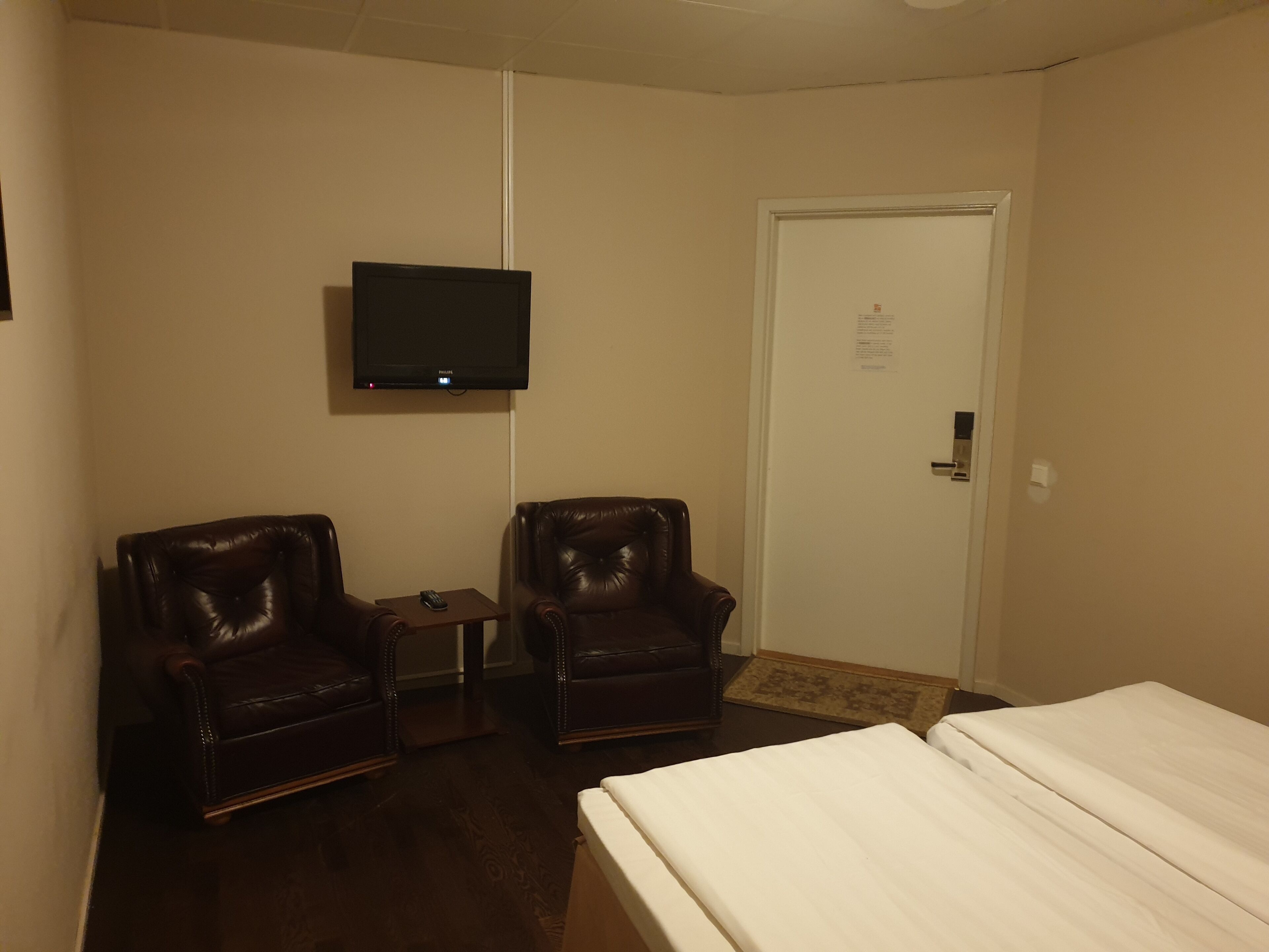 Une chambre d’hôtel avec deux fauteuils en cuir, un lit, un téléviseur fixé au mur et une porte avec une pancarte.
