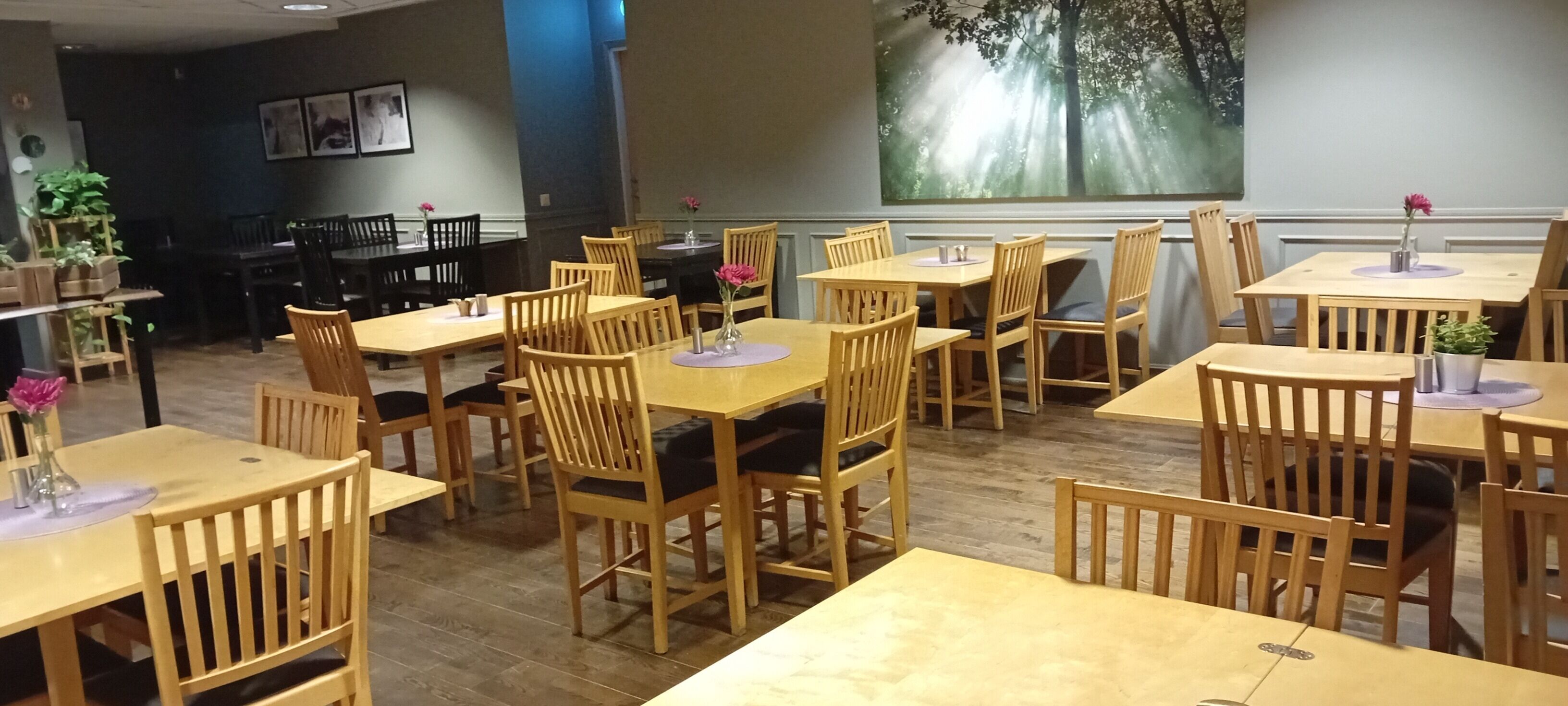 Ein Restaurant mit Holztischen und -stühlen, einem großen Gemälde an der Wand und Kübelpflanzen.