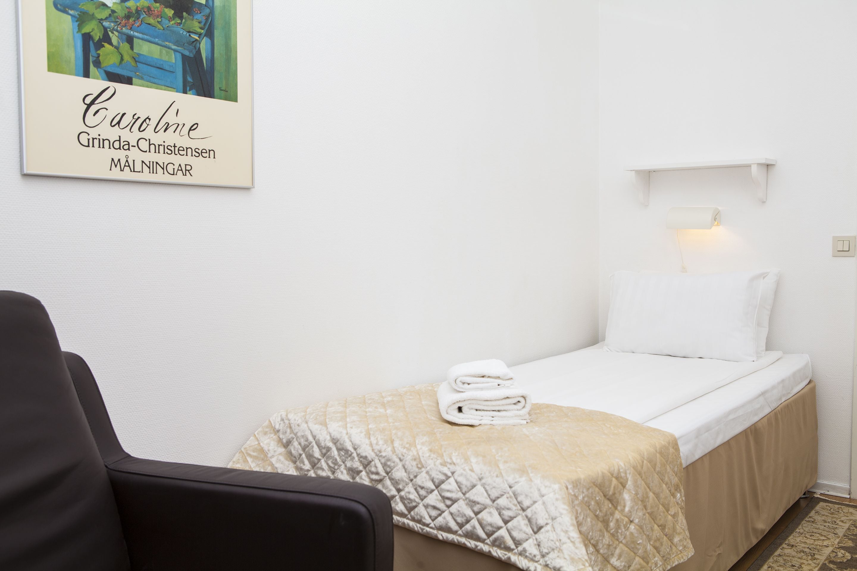 A small, neatly arranged bedroom with a bed, a chair, a quilted blanket, and a wall-mounted shelf with a lamp.