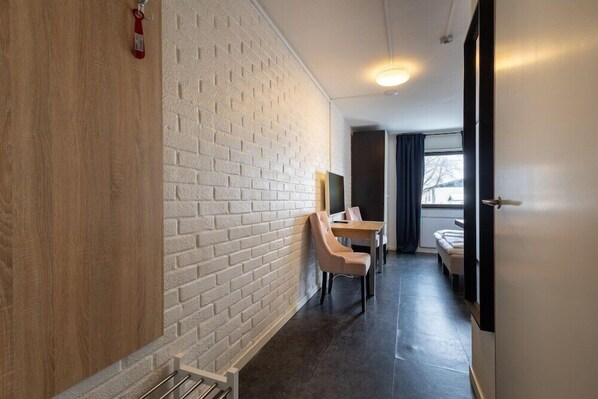 A hallway with a white brick wall, a wooden panel, a desk with a chair, a bed, and a door. - Arena Hotel (Halmstad)