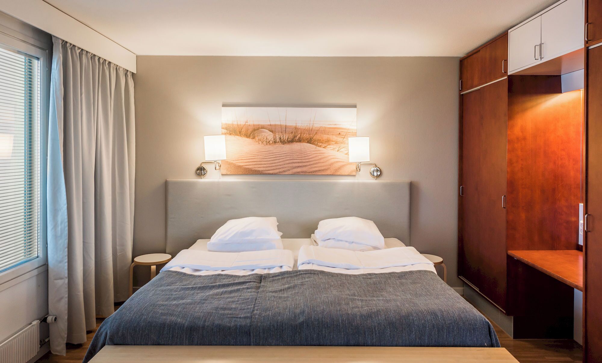 Moderni hotellihuone, jossa on suuri sänky, kaksi yöpöytää, yölamppu, seinään kiinnitetty valaisin ja suuri taideteos sängyn yläpuolella.