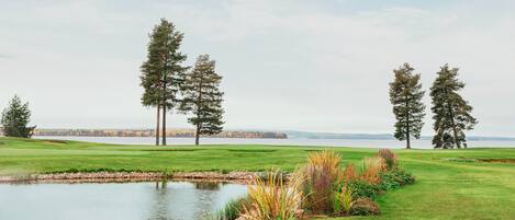 A golf course with a pond, trees, and a view of a distant lake.