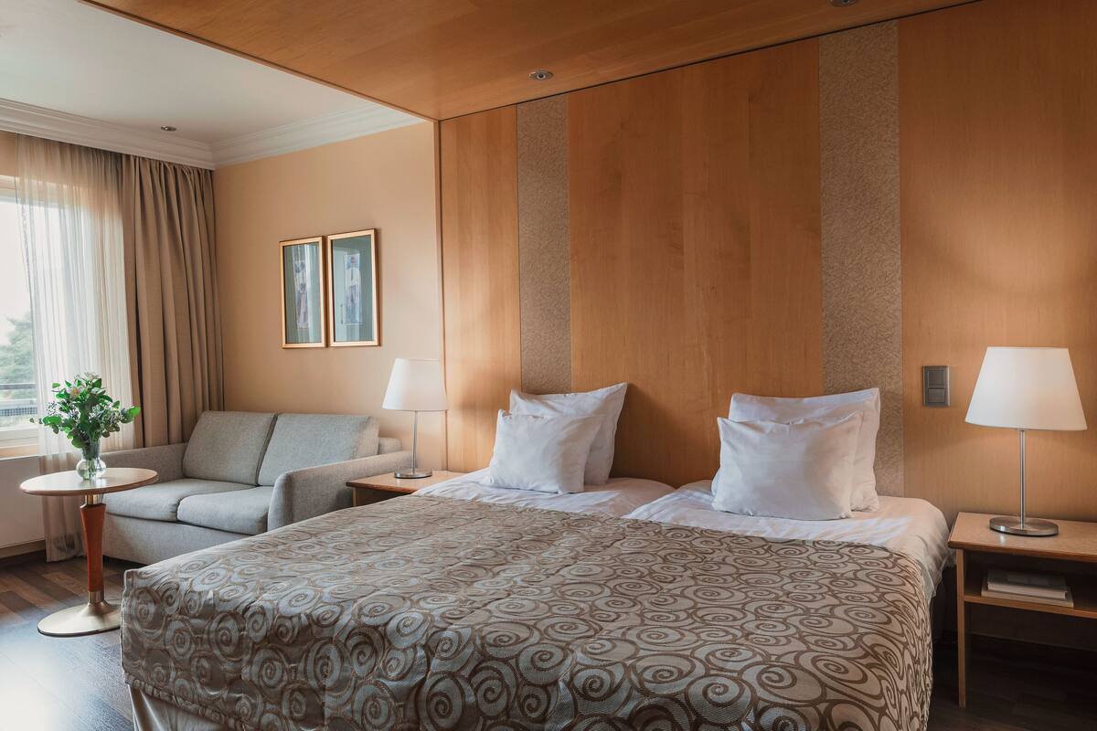 A hotel room with a bed, a sofa, two bedside tables, a lamp, a small table with a vase, and a window with curtains.