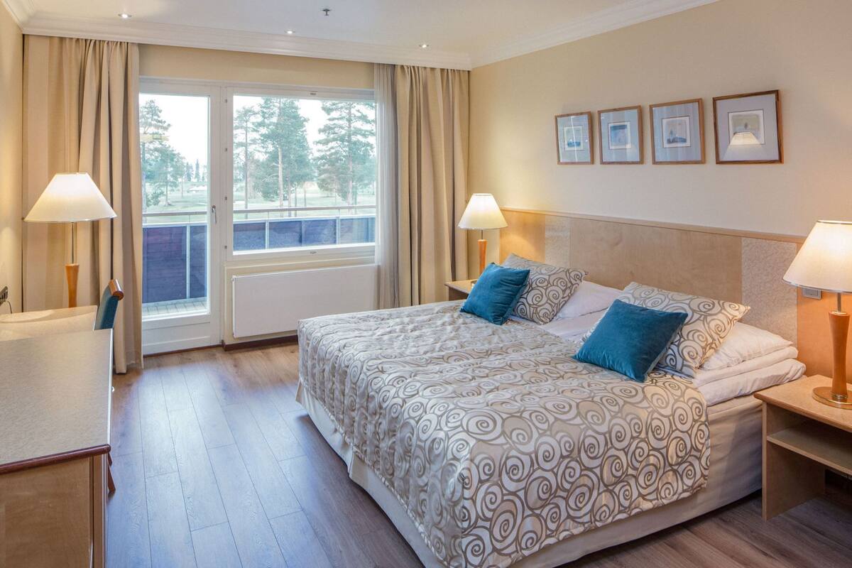 A hotel room with a bed, two bedside lamps, a window with curtains, a radiator, and framed pictures on the wall.