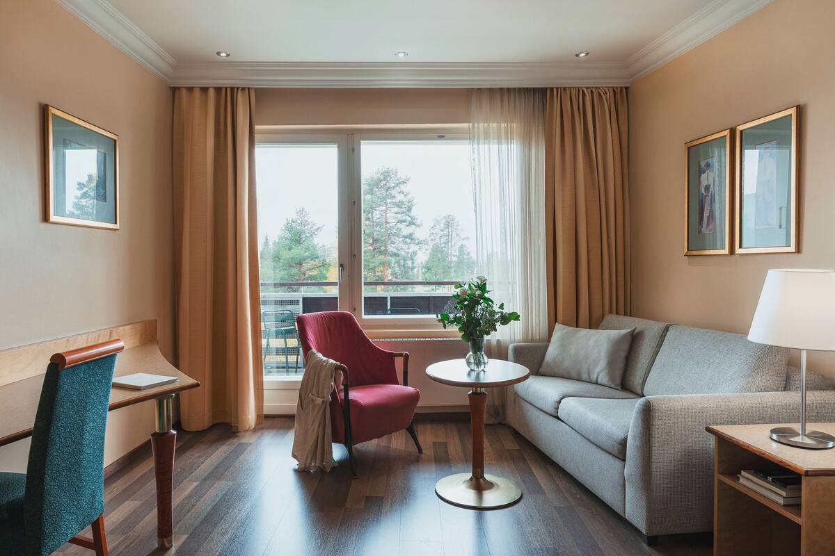 A hotel room with a red armchair, a grey sofa, a small table with a vase, and a lamp.