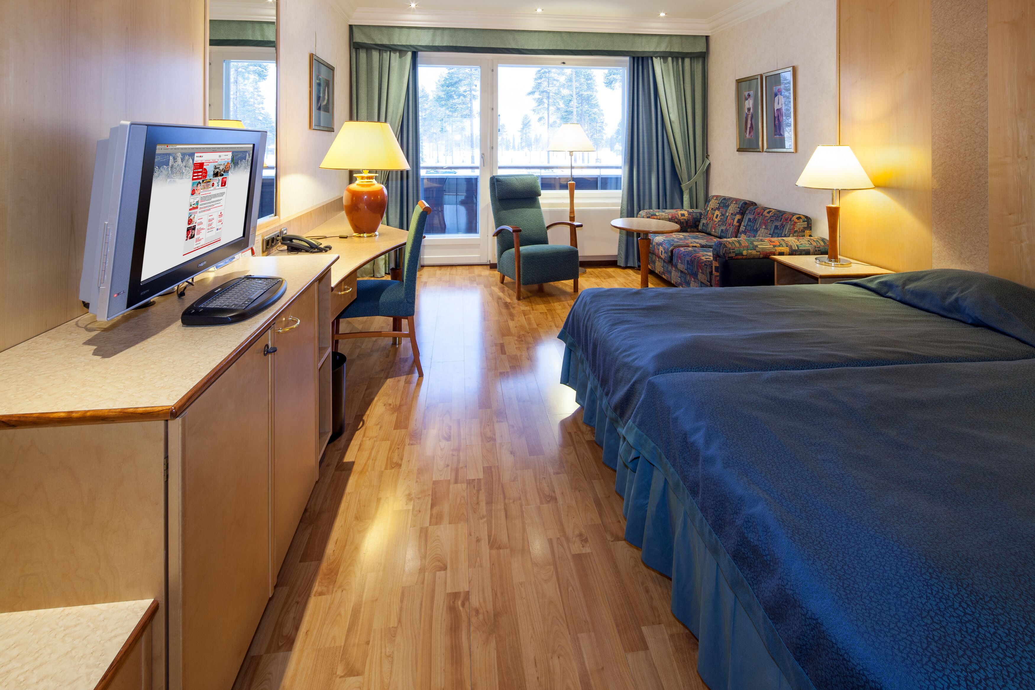 Habitación de hotel con cama, escritorio, computadora, silla y una pequeña mesa con lámpara.