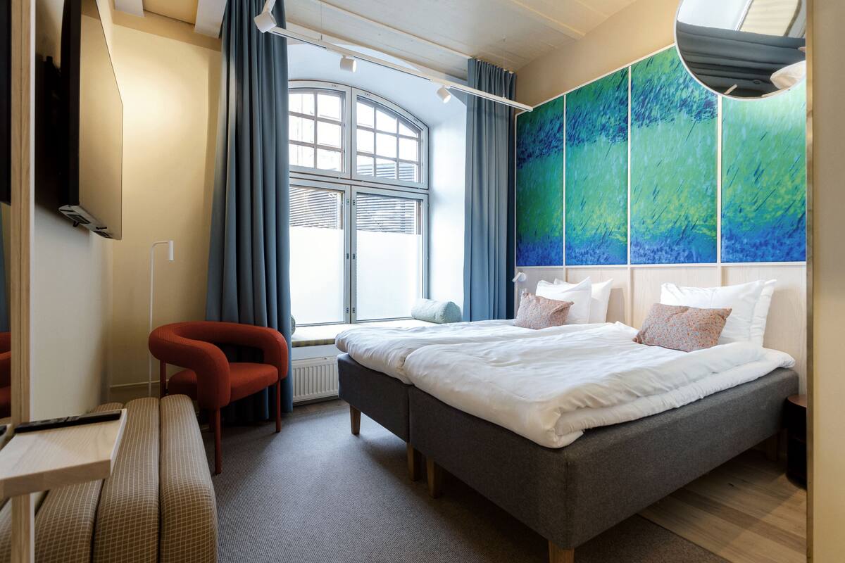 A modern hotel room with a large bed, a desk, a chair, and a large window with curtains.