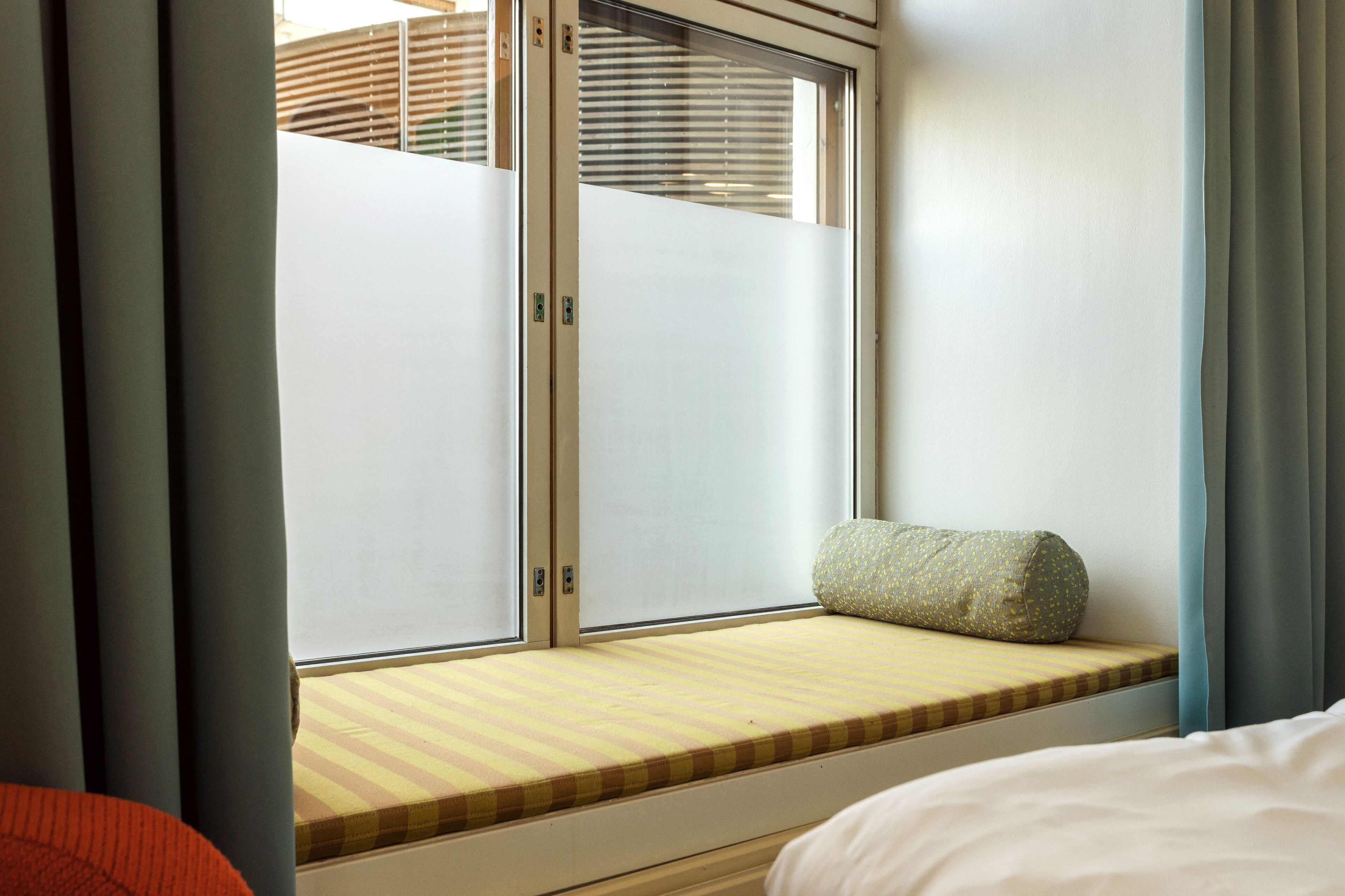 Una habitación con cama, una ventana con persianas y un pequeño banco con cojín.