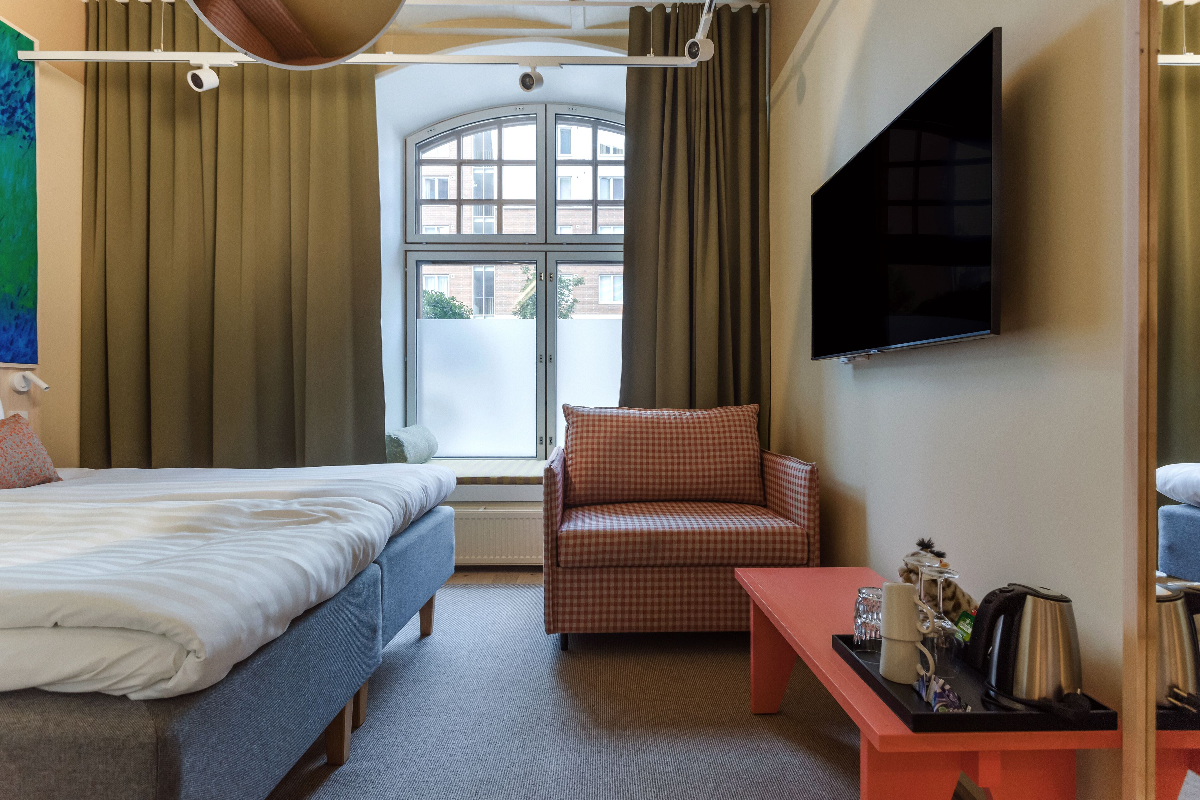 A hotel room with a bed, a checkered armchair, a small table with a kettle and cups, and a TV mounted on the wall.