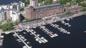 Vista aérea de un puerto deportivo con numerosas embarcaciones amarradas, rodeado de edificios residenciales y un gran edificio de ladrillo con tejado inclinado.
