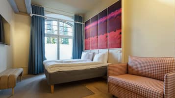 A bedroom with a bed, a bench, a sofa, a large window with curtains, and a wall with a large painting.