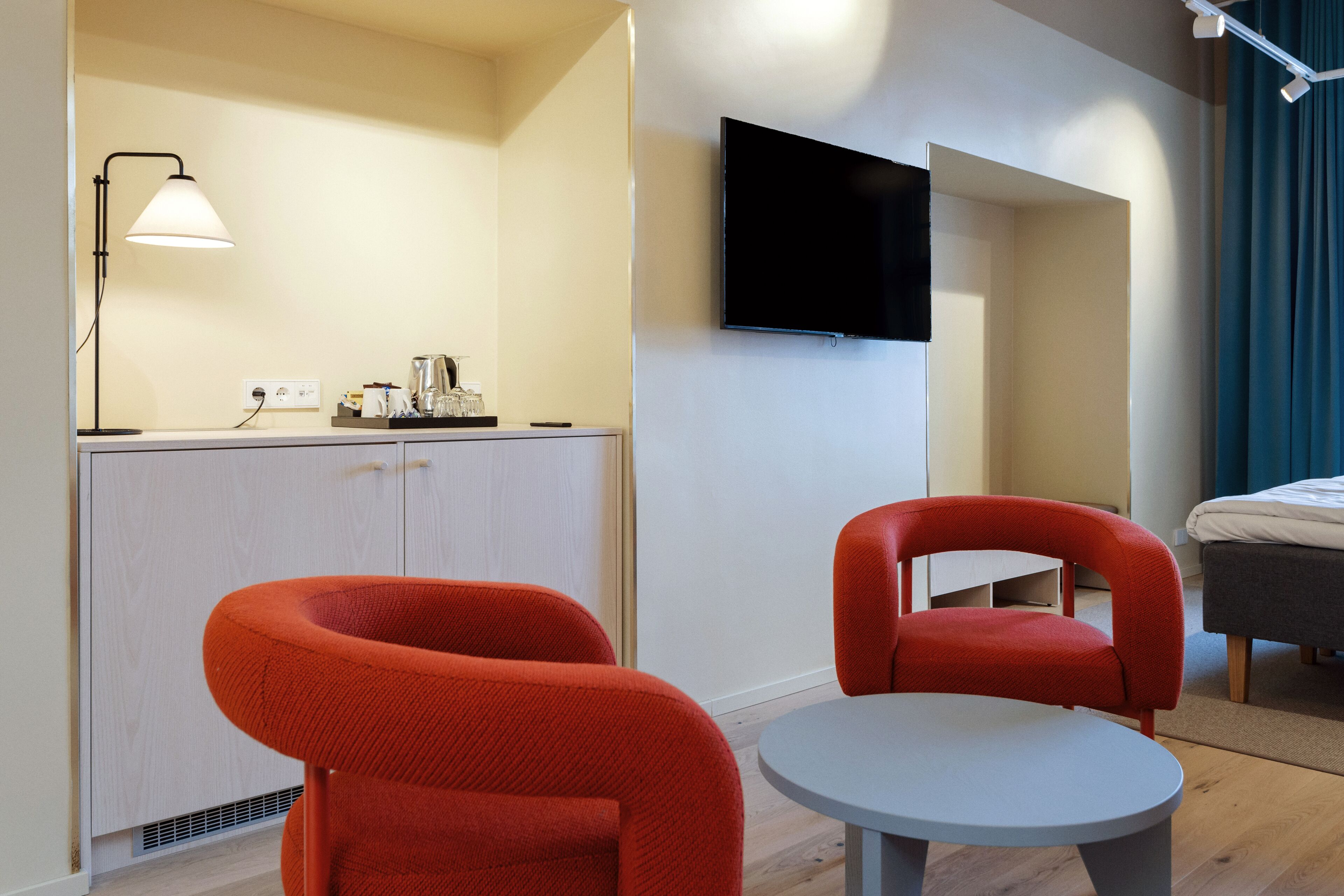 Une chambre d’hôtel moderne équipée d’un téléviseur à écran plat fixé au mur, d’une petite table ronde, de deux chaises rouges, d’un lit avec une tête de lit grise et d’un bureau avec une lampe et une machine à café.