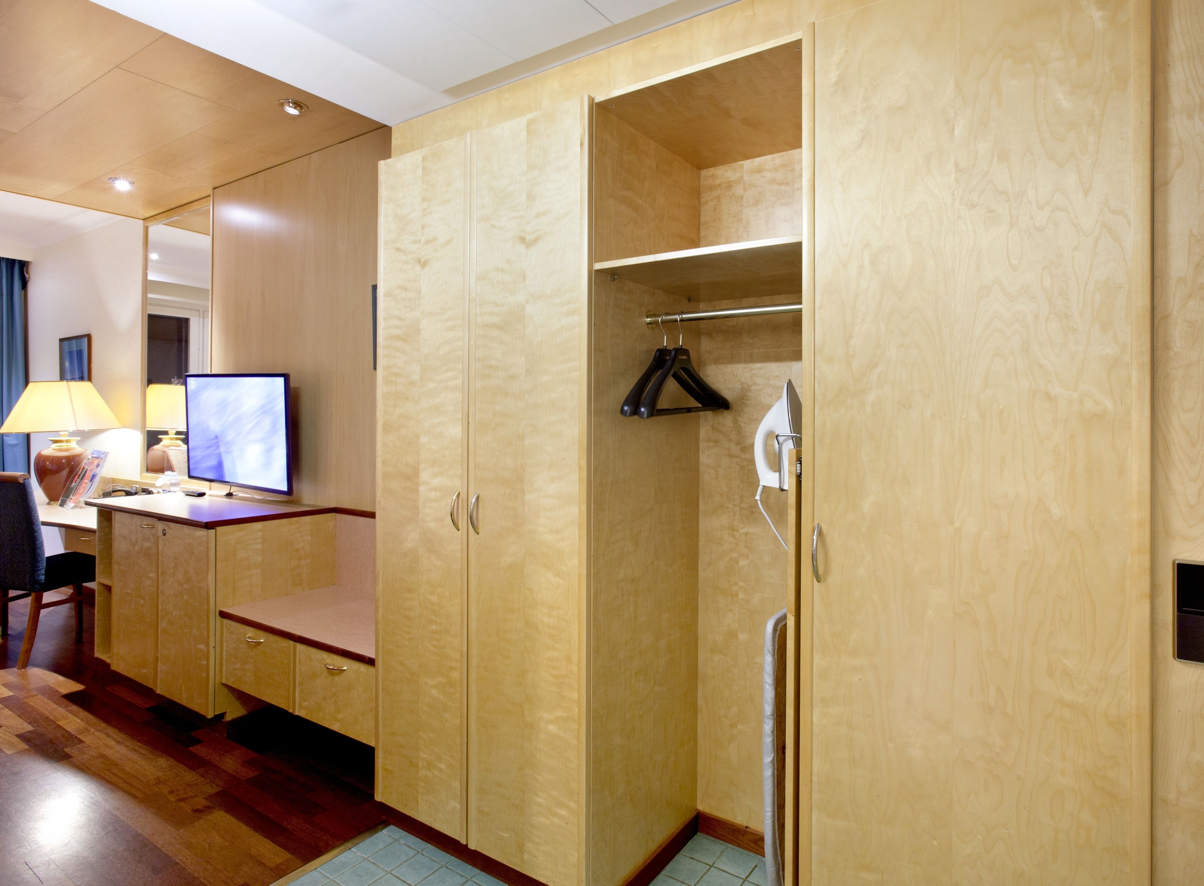 Una habitación de hotel con un armario de madera, un escritorio con computadora, una lámpara y una silla.