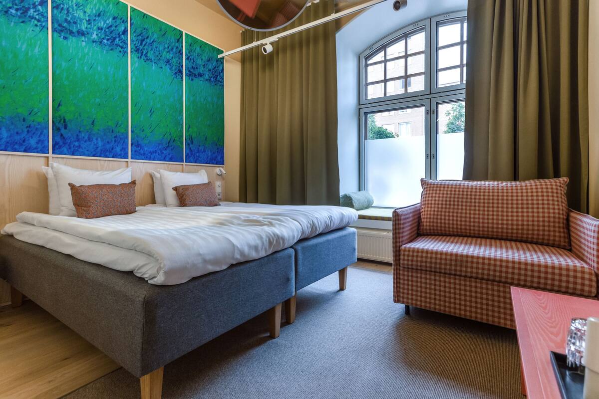 A modern hotel room with a large bed, a sofa, a window with curtains, and a colorful abstract painting on the wall.