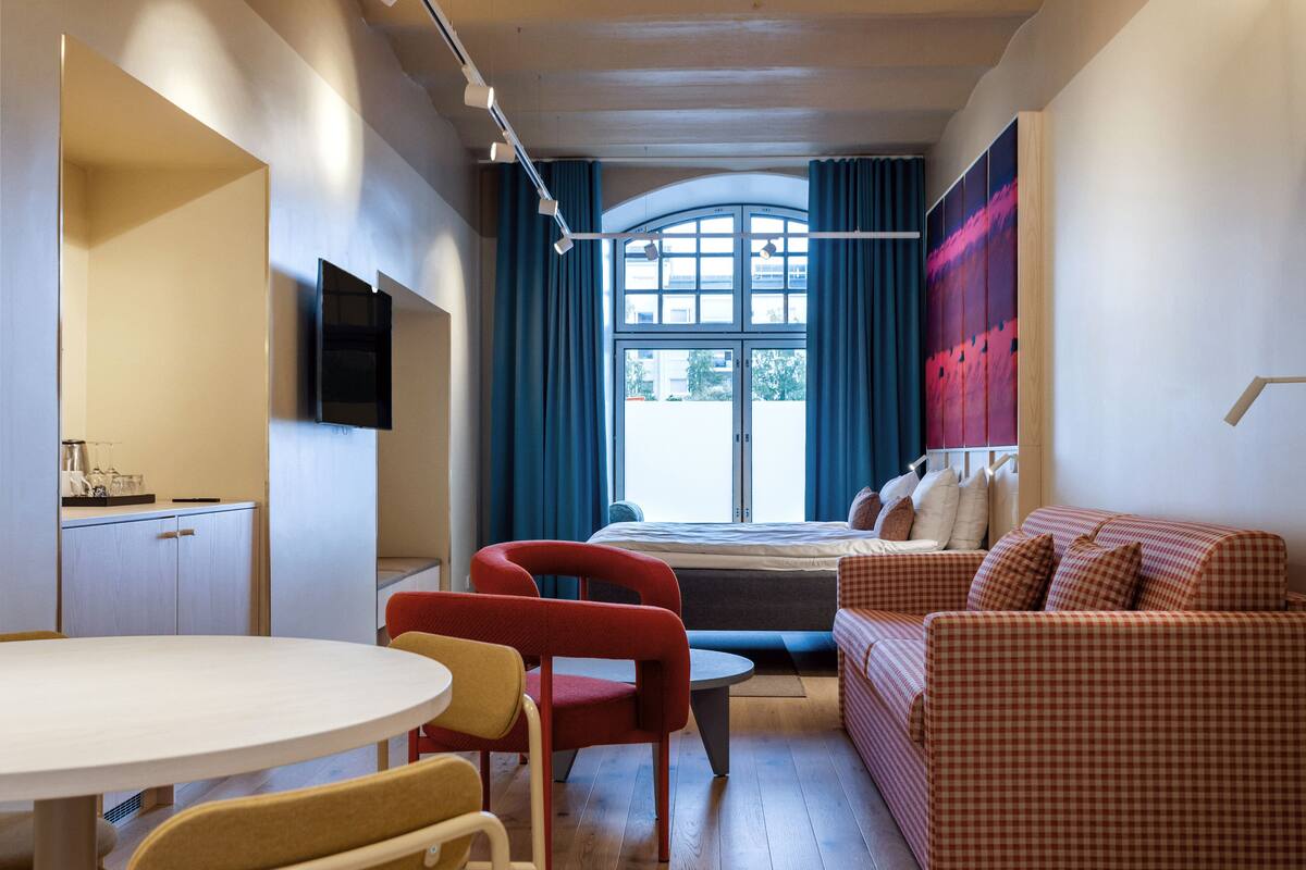 A compact room with a bed, a red and white checkered sofa, a small table, and a TV mounted on the wall.