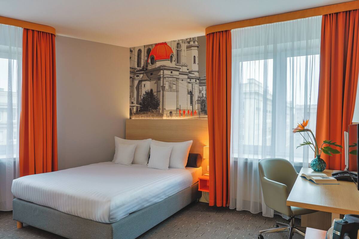 A hotel room with a bed, a desk with a chair, a lamp, and a painting on the wall.