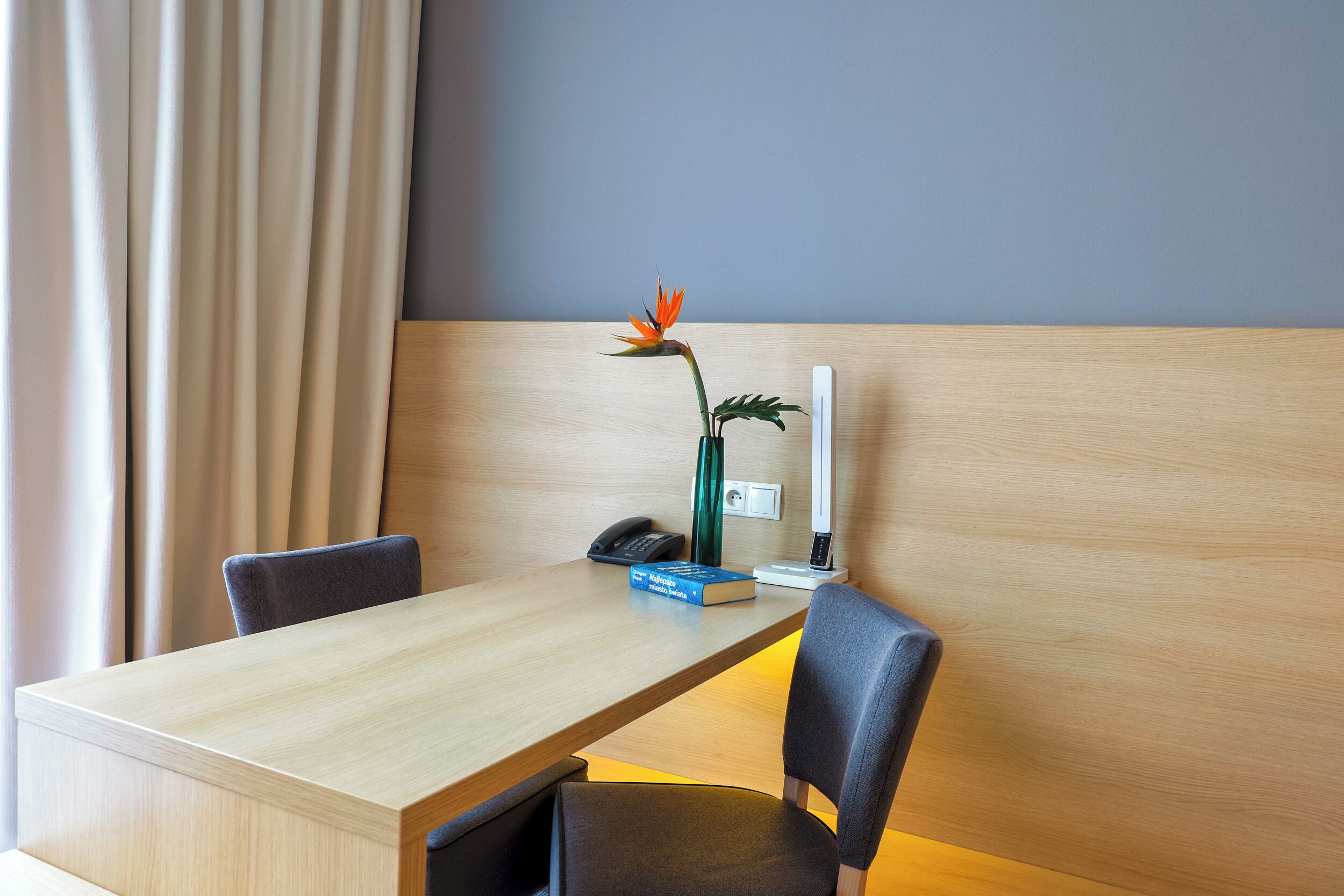 A hotel room with a wooden desk, two chairs, a vase with a flower, a book, a phone, and a bedside table with a lamp.