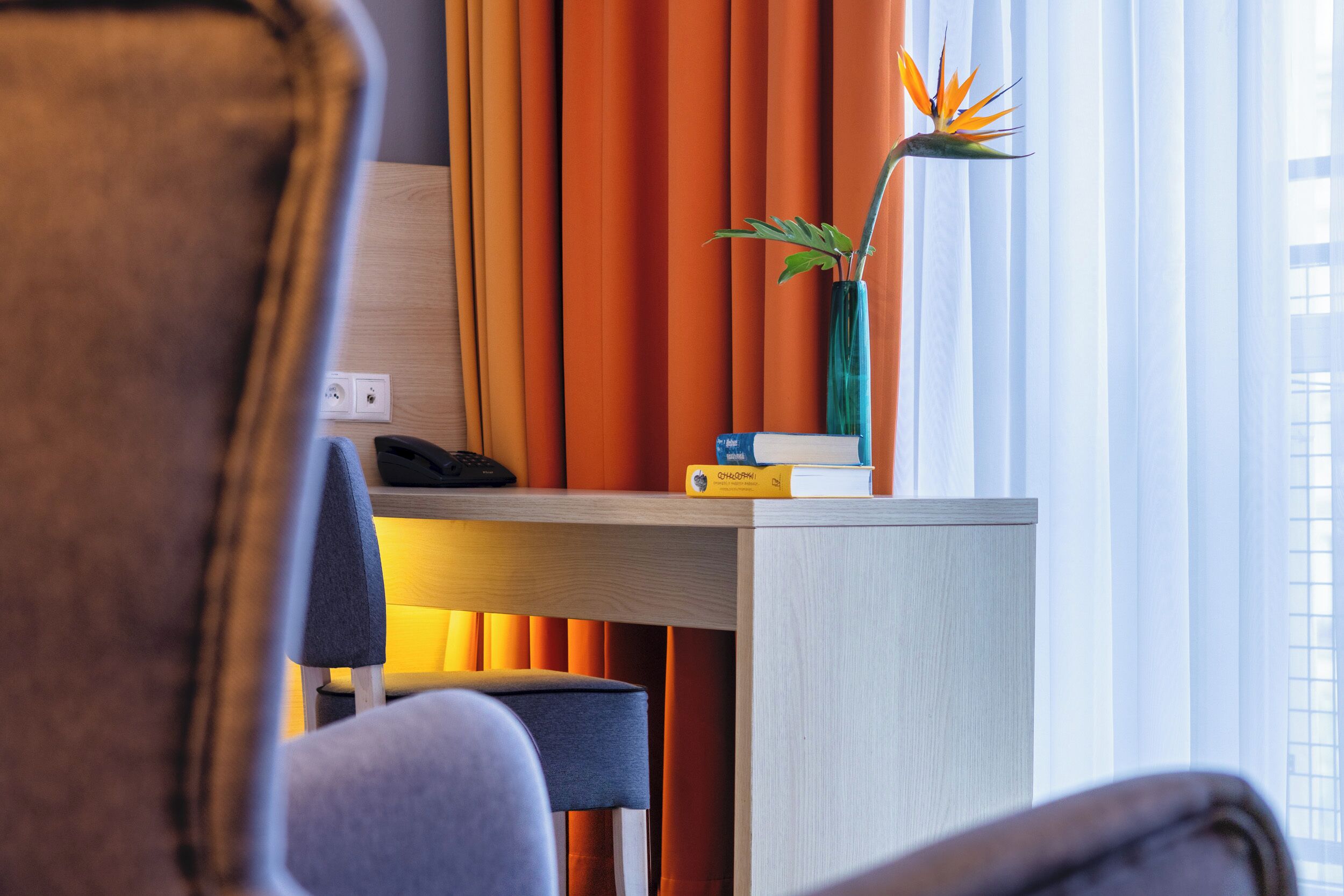 Habitación de hotel con escritorio, silla, jarra con planta y cortinas naranjas.