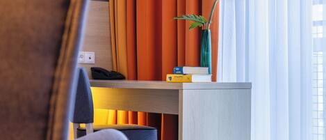 Habitación de hotel con escritorio, silla, jarra con planta y cortinas naranjas.