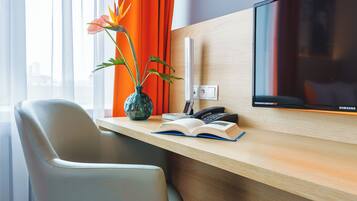 A hotel room with a wooden desk, a modern TV, a vase with flowers, and a city view.